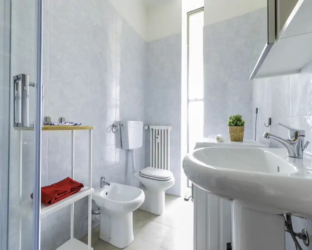 Baño moderno y luminoso con lavabo, bidé y inodoro; azulejos claros y una ventana alta proporcionan abundante luz natural.