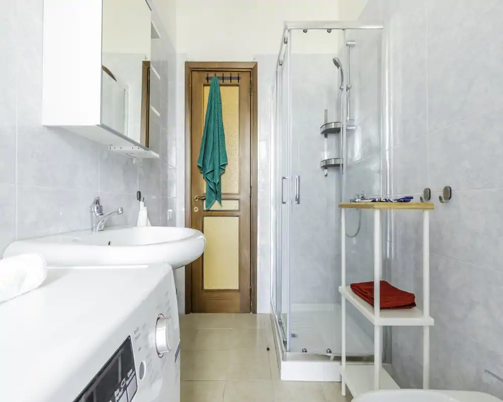 Baño luminoso y limpio con cabina de ducha de cristal, lavabo y lavadora visibles. Baldosas neutras y estanterías sencillas que hacen el espacio práctico y bien iluminado.