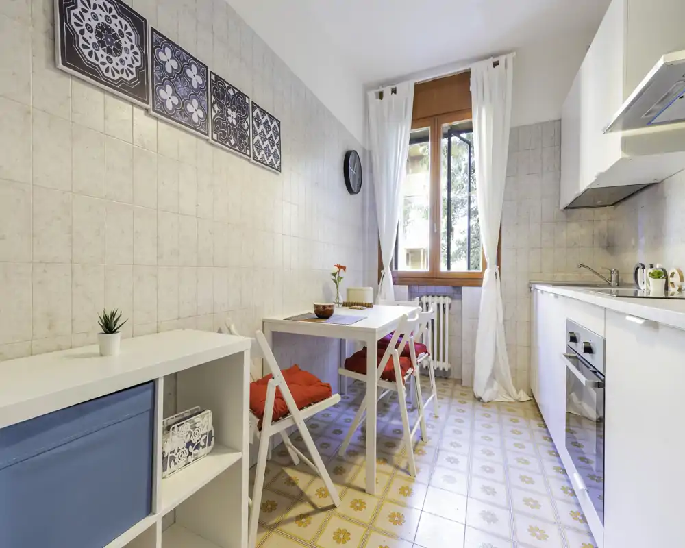 Cocina luminosa y compacta con mesa pequeña, sillas con cojines rojos, suelos de baldosa decorativa y mobiliario blanco — ideal para comidas acogedoras en la ciudad.