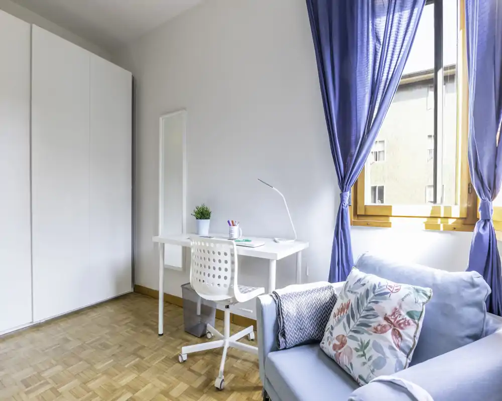 Angolo luminoso e ordinato in stile camera da letto con una scrivania, armadio e una seduta imbottita che può fungere da posto letto. La luce naturale dalla finestra e i tessili morbidi creano un ambiente fresco e accogliente.