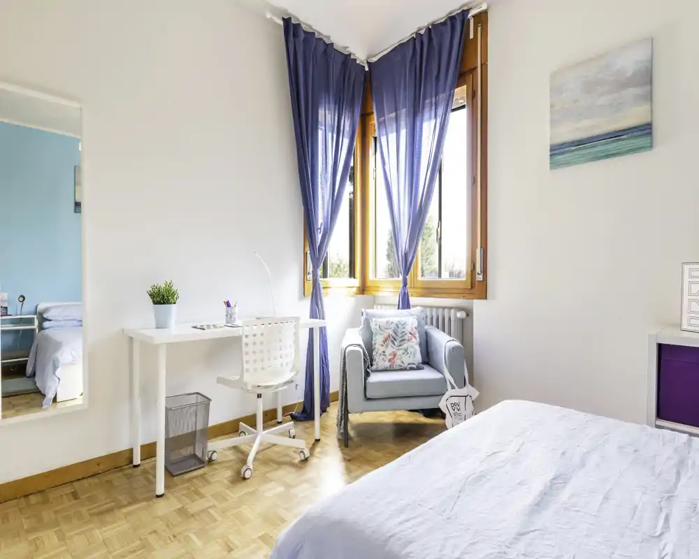 Camera da letto luminosa e ariosa con letto in primo piano, una piccola scrivania e una poltrona accogliente vicino alla finestra illuminata, con tende blu.