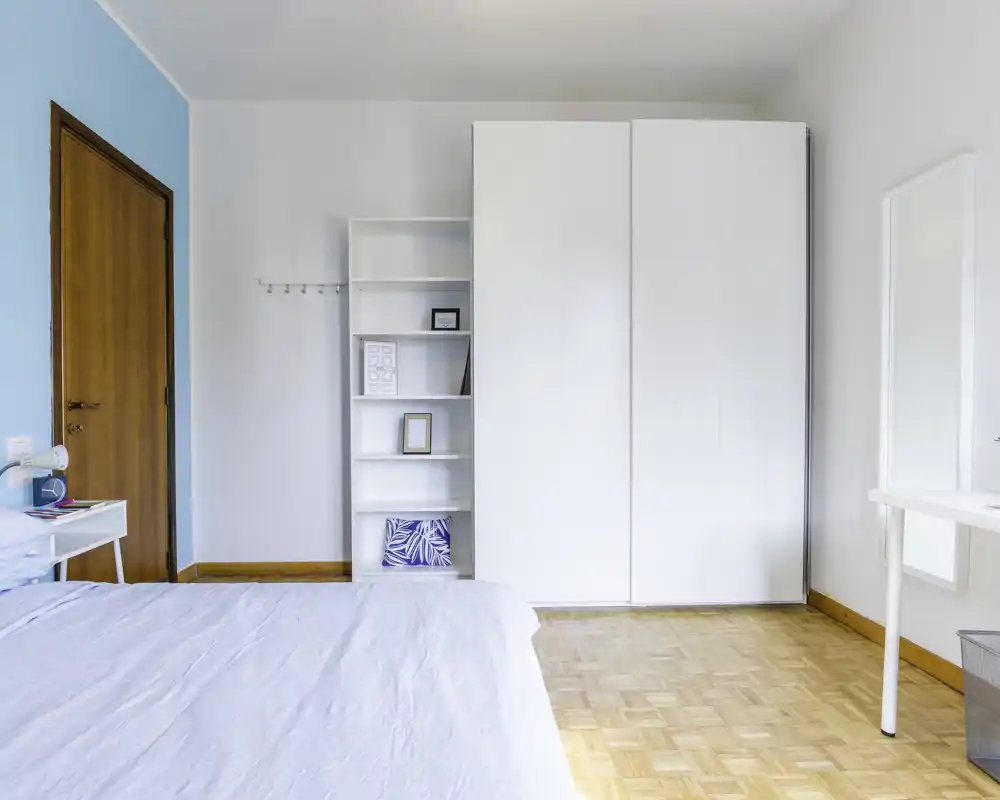 Camera da letto luminosa e minimal con letto ben rifatto, grande armadio bianco e una scrivania semplice. Pavimento in legno e luce naturale creano un'atmosfera ariosa e tranquilla, perfetta per annunci.
