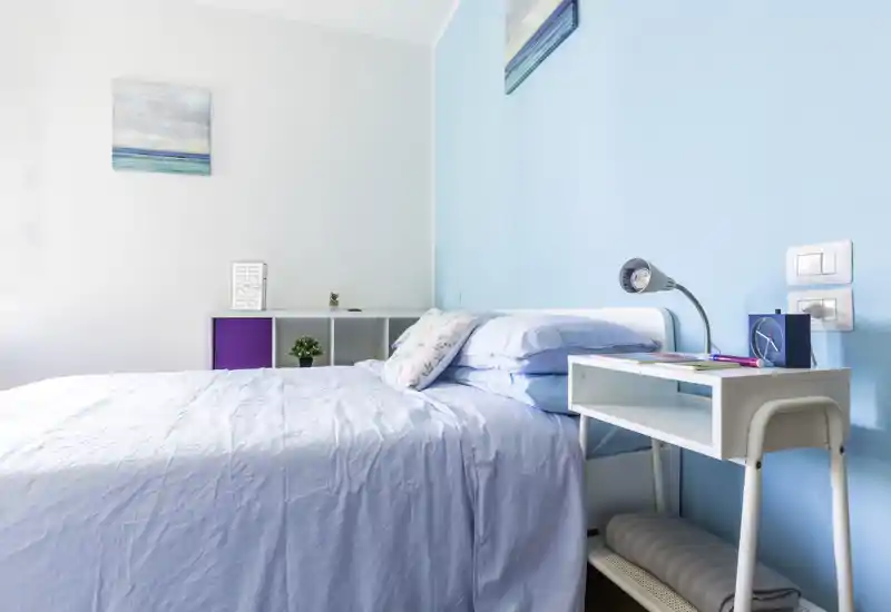 Camera da letto luminosa e moderna con pareti azzurro chiaro e letto matrimoniale ben rifatto. Piccolo scrittoio e mensole minimal rendono l'ambiente arioso e adatto agli annunci.