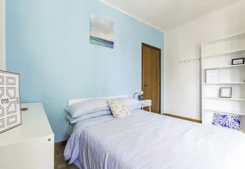 Camera da letto luminosa e ariosa con letto matrimoniale con biancheria azzurro chiaro, cuscino decorativo e mensole minimal; parete accent turchese e luce naturale creano un'atmosfera rilassante.