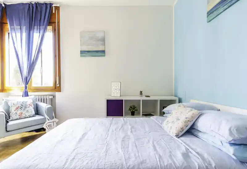 Camera da letto luminosa e ariosa con un ampio letto con biancheria chiara, una poltrona accogliente vicino alla finestra e dettagli dallo stile marino per un'atmosfera rilassata.