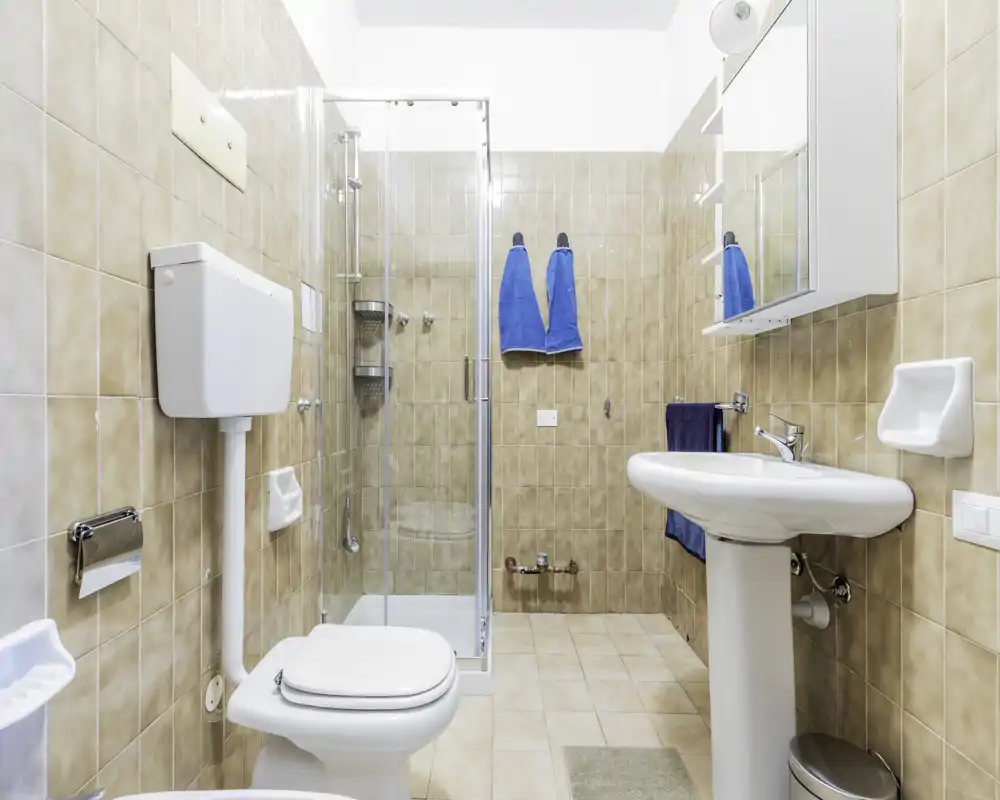Bagno luminoso e pulito con box doccia, lavabo a colonna, WC e pensile specchiato. Piastrelle beige neutro e asciugamani blu creano un piacevole contrasto.