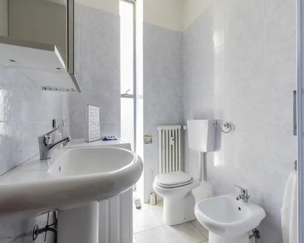 Bagno luminoso e pulito con lavabo ampio, WC e bidet; piastrelle chiare e luce naturale da una finestra alta rendono l'ambiente fresco.