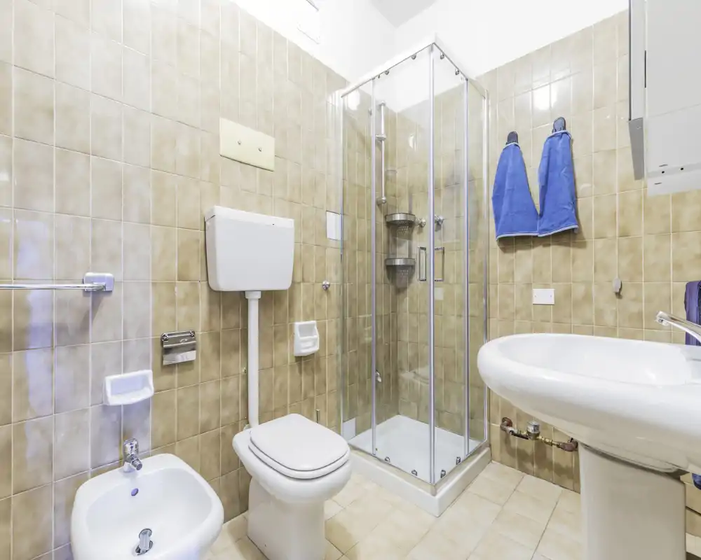 Bagno pulito e ben illuminato con cabina doccia angolare in vetro, WC, bidet e lavabo a colonna; piastrelle beige neutre e luce brillante.