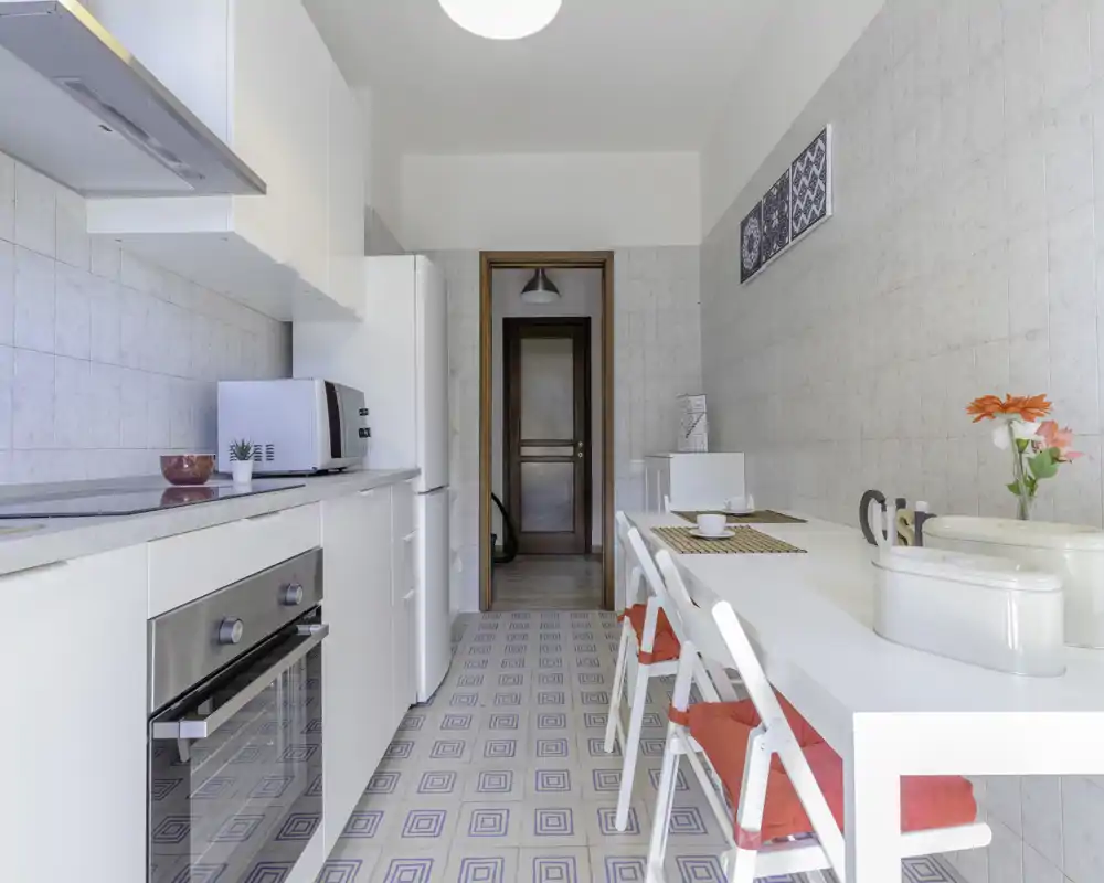 Cucina luminosa e moderna con mobili bianchi, forno incassato, frigorifero e un tavolo da pranzo con sedie. Stile pulito e dettagli decorativi come i cuscini rossi e i fiori freschi creano un'area pranzo accogliente.