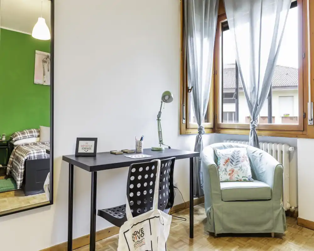 Coin chambre lumineux et soigné avec un bureau et un fauteuil confortable près d'une grande fenêtre ; lit visible dans une alcôve adjacente avec des textiles chaleureux.