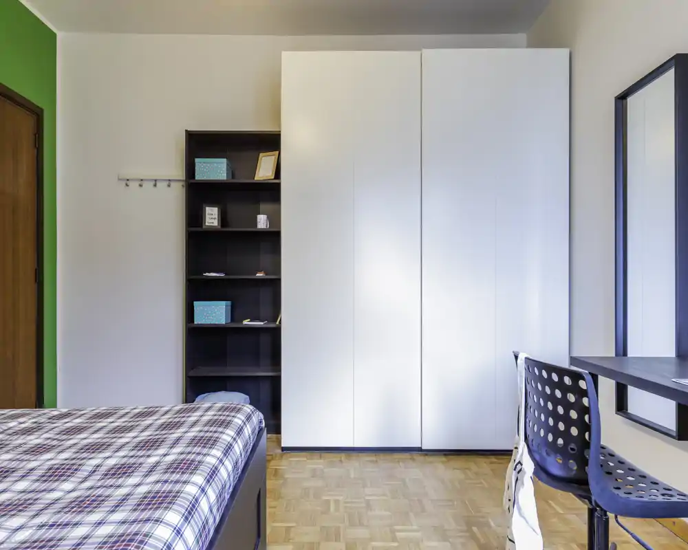 Chambre lumineuse et bien rangée avec lit à carreaux, grand placard blanc et bureau près de la fenêtre — parfaite pour étudiant ou locataire seul.