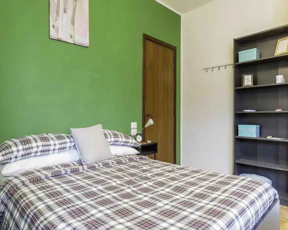 Chambre lumineuse et confortable avec un grand lit à carreaux devant un mur vert, table de chevet avec lampe et étagère ouverte pour le rangement.