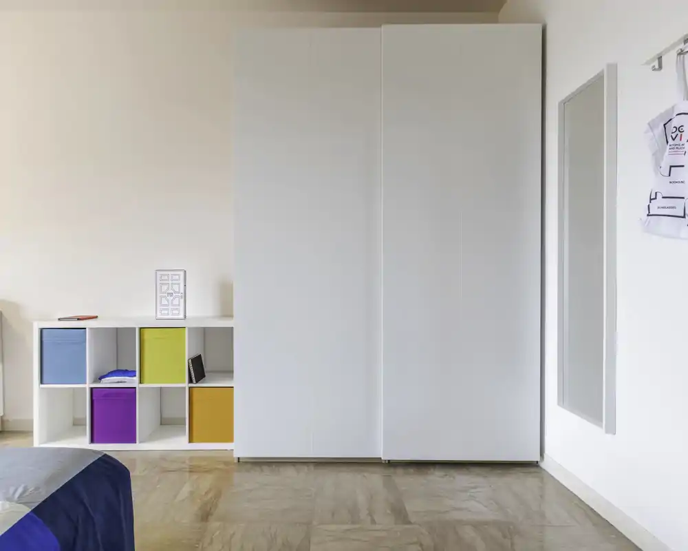 Angolo di camera luminoso e minimal con un grande armadio bianco, contenitori a cubi colorati e vista parziale del letto in primo piano. Linee pulite e luce naturale rendono l'ambiente fresco e arioso, ottimo per gli annunci.
