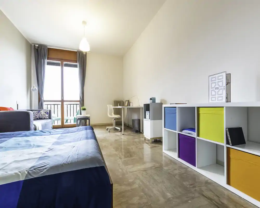 Camera da letto luminosa e moderna con letto matrimoniale visibile, una scrivania vicino alla finestra e contenitori a cubo colorati lungo la parete; grandi porte sul balcone offrono molta luce naturale.