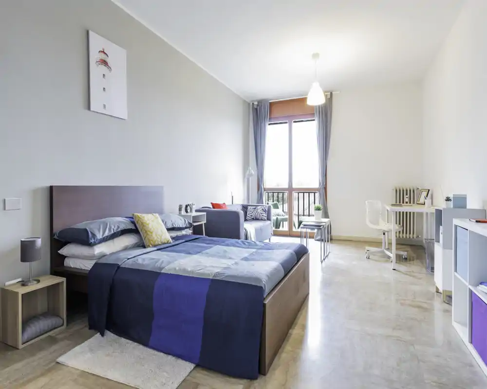 Camera da letto luminosa e moderna con letto matrimoniale in primo piano, tessili accoglienti e un piccolo angolo salotto vicino al balcone con luce naturale.