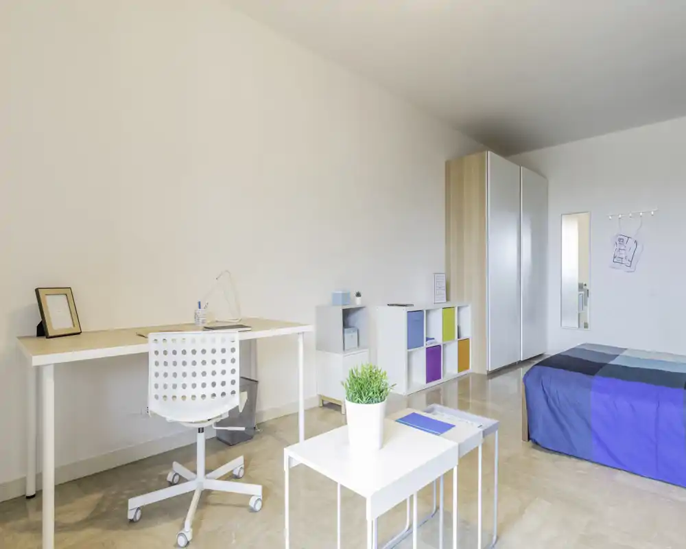 Camera da letto luminosa e moderna con letto singolo blu, scrivania con sedia ergonomica e scaffalature minimaliste—ideale per uno studente o una persona sola.
