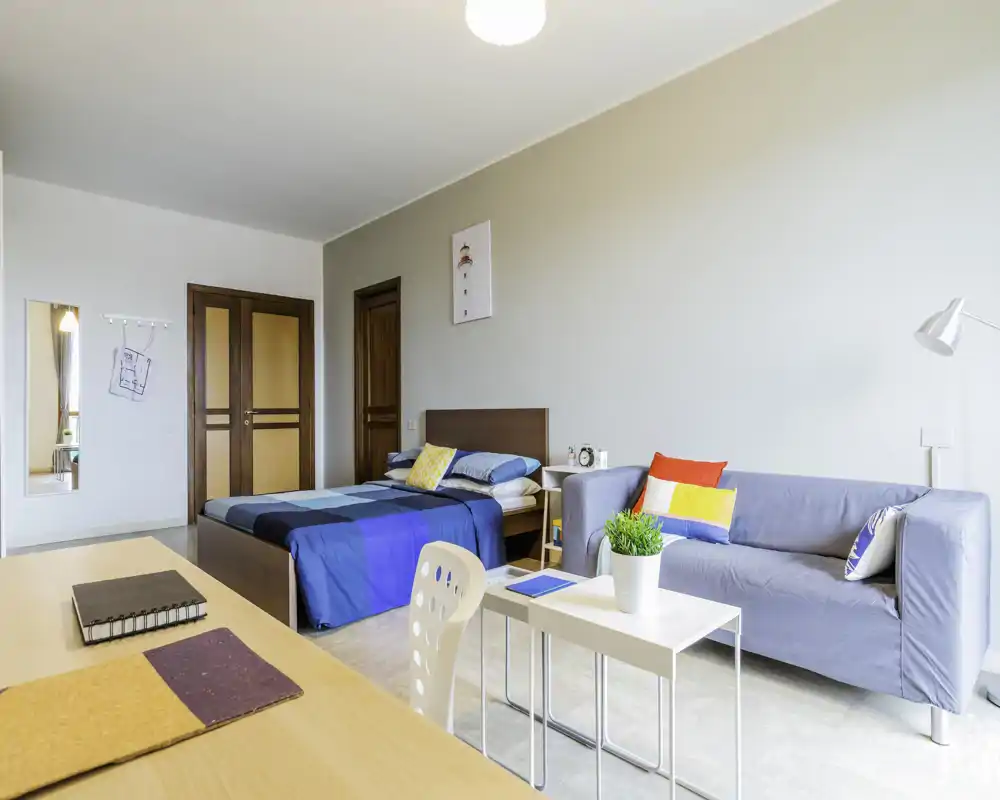 Camera da letto luminosa e moderna con letto matrimoniale, comodino e una piccola zona soggiorno con divano e tavolino. Stile pulito, tessuti colorati e buona luce naturale la rendono accogliente.