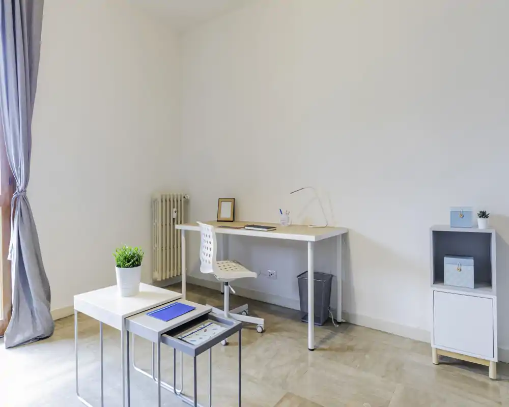 Zona giorno minimalista e luminosa con scrivania semplice, sedia e tavolini annidati; luce naturale intensa e arredo moderno e pulito, ideale per il lavoro da casa.