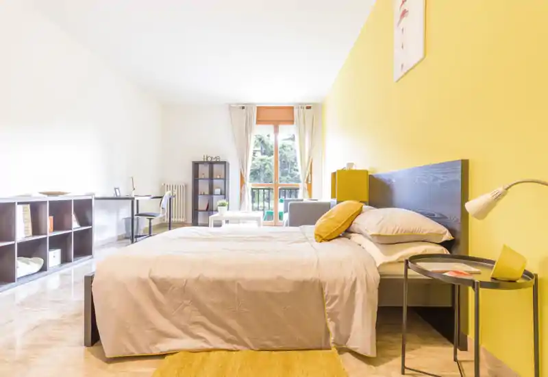 Dormitorio moderno y luminoso con cama doble bien tendida, pared amarilla y luz natural desde el balcón. Estanterías ordenadas, zona de trabajo y textiles suaves proporcionan un espacio cómodo y bien decorado, perfecto para alquileres temporales.