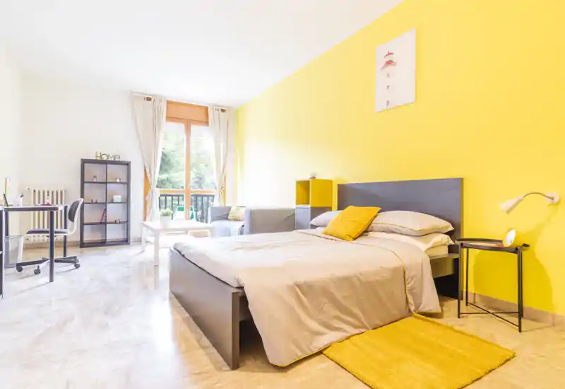 Dormitorio moderno y luminoso con cama doble, pared de acento amarilla y mucha luz natural por una ventana grande. La habitación cuenta con mesita, escritorio y una pequeña zona de estar — perfecta para una persona o pareja.