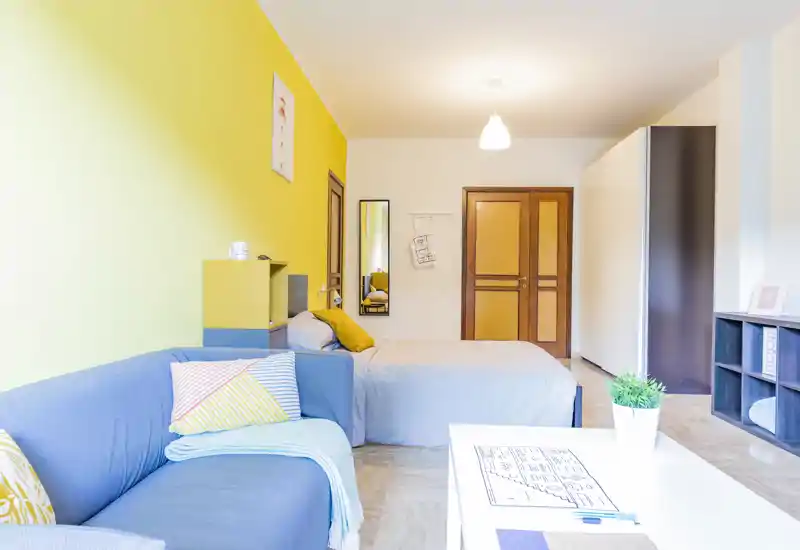 Camera luminosa e moderna con letto matrimoniale, divano blu e parete accentuata gialla. Spazio ben illuminato e arredato con gusto, cuscini e tavolino, ideale per affitti brevi.
