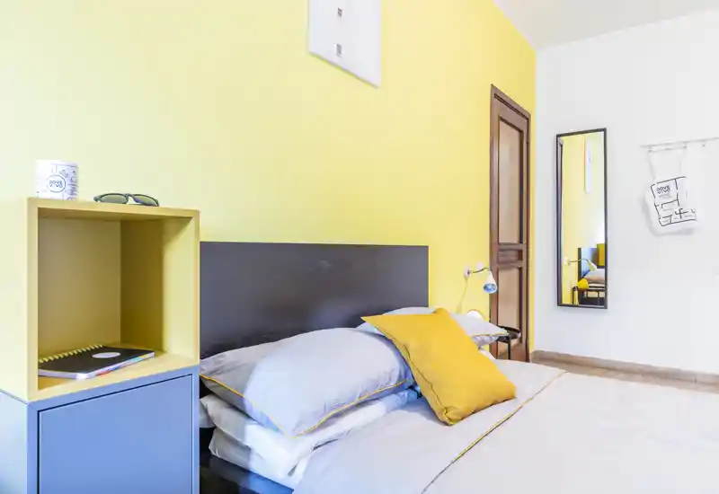 Dormitorio moderno y luminoso con pared amarilla, cama bien hecha con ropa gris y cojín amarillo; estantería minimalista y espejo de cuerpo entero completan la escena.