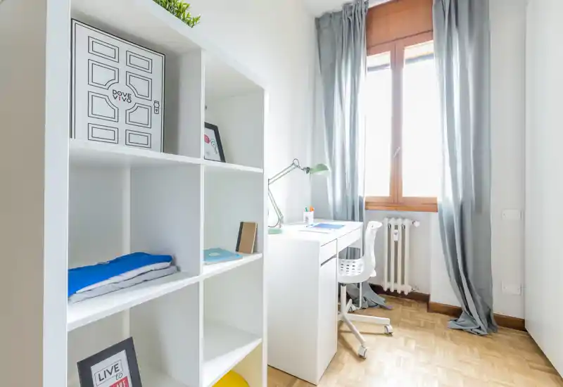 Coin chambre compact et lumineux avec une étagère blanche et un petit bureau près d'une fenêtre ensoleillée ; parquet clair et rideaux gris apportent une ambiance propre et apaisante.