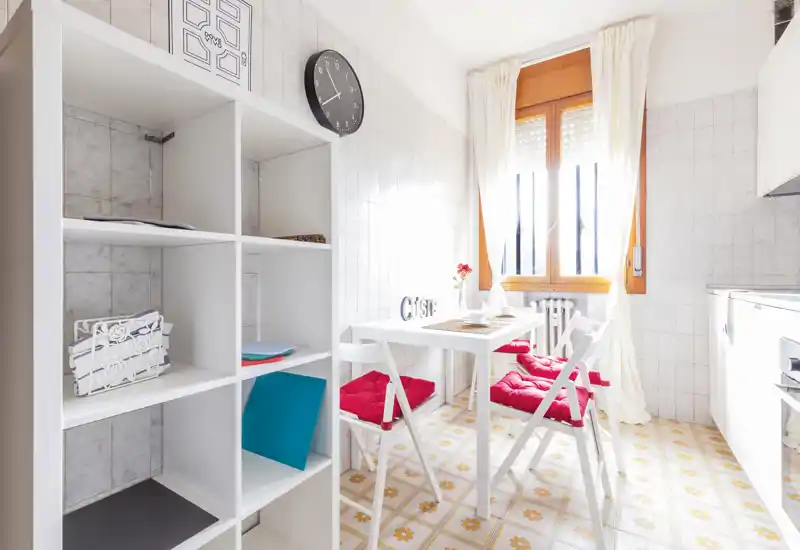 Bright, compact kitchen with a small dining table and red-cushioned folding chairs, white shelving and tiled floor—ideal for a cozy city apartment.