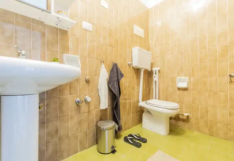 Compact, clean bathroom with a pedestal sink, toilet with high cistern, towel hooks and a small trash can. Warm beige tiles and bright lighting create a tidy, functional space.