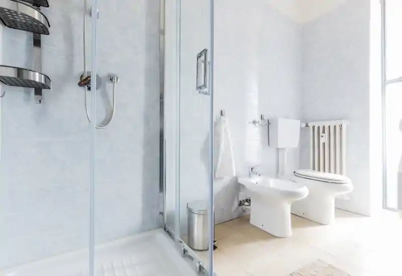 Bright, clean bathroom with a glass shower enclosure, bidet and toilet, light tiled walls and a window providing natural light.