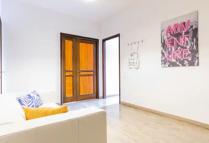 Bright, modern living area with a white sofa accented by colorful cushions, clean floors and a wooden entrance door; minimal, well-lit space ideal for a rental listing.