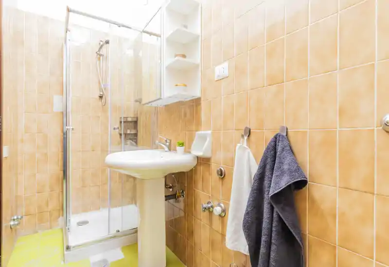 Clean, compact bathroom with a corner shower, pedestal sink and wall-mounted shelving. Warm terracotta tiles and bright lighting create an inviting, well-maintained space ideal for short-term rentals.