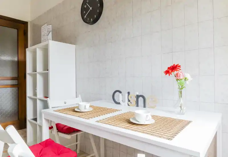 Bright, compact kitchen dining area with a white table set for two, decorative placemats and a vase with flowers creating a pleasant, well-styled space.