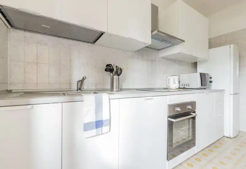 Bright, modern compact kitchen with white cabinetry, built-in oven, fridge and countertop appliances; tidy minimalist styling ideal for a rental listing.