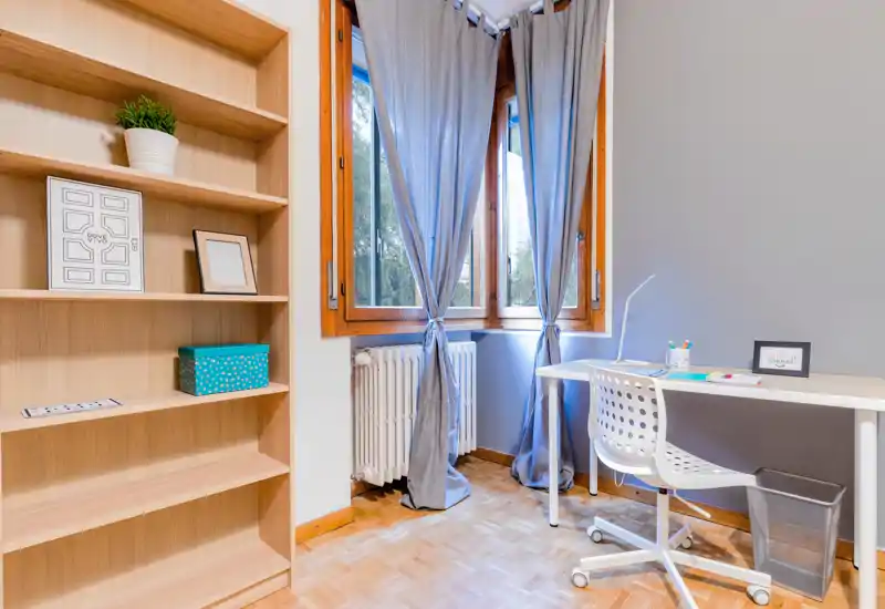 Bright, tidy home workspace with a white desk and ergonomic chair next to large windows dressed with gray curtains; built-in shelving and wooden parquet floor add warm, functional character.