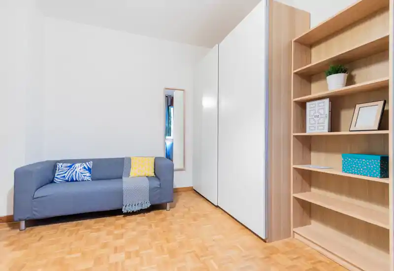 Bright, minimal living area with a compact blue sofa, decorative cushions and a wooden shelving unit providing storage and display space.
