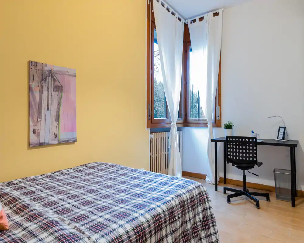 Bright, tidy bedroom with a double bed dressed in a checked duvet, a window with sheer curtains providing natural light, and a simple work desk beside the window — comfortable and uncluttered space ideal for short-stay rentals.