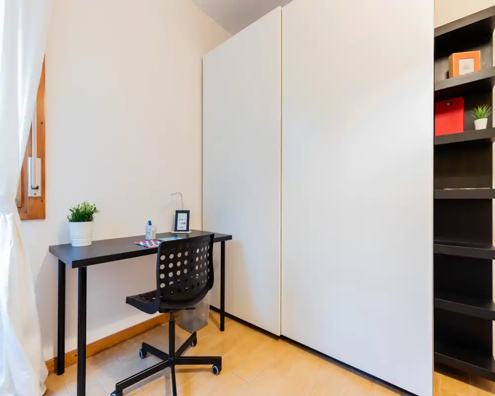 Compact bedroom corner set up as a study area with a simple desk, swivel chair and built-in wardrobe; bright natural light and clean, minimal styling.