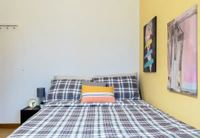 Bright, neatly styled bedroom with a full-size bed dressed in patterned plaid linens, accent pillows and modern wall art on a yellow accent wall.