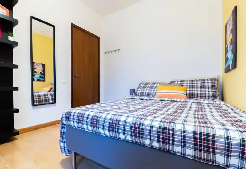 Bright, neatly made bedroom with a patterned double bed, decorative pillows and a full-length mirror. Clean lines and warm wooden flooring create a welcoming, modern feel.