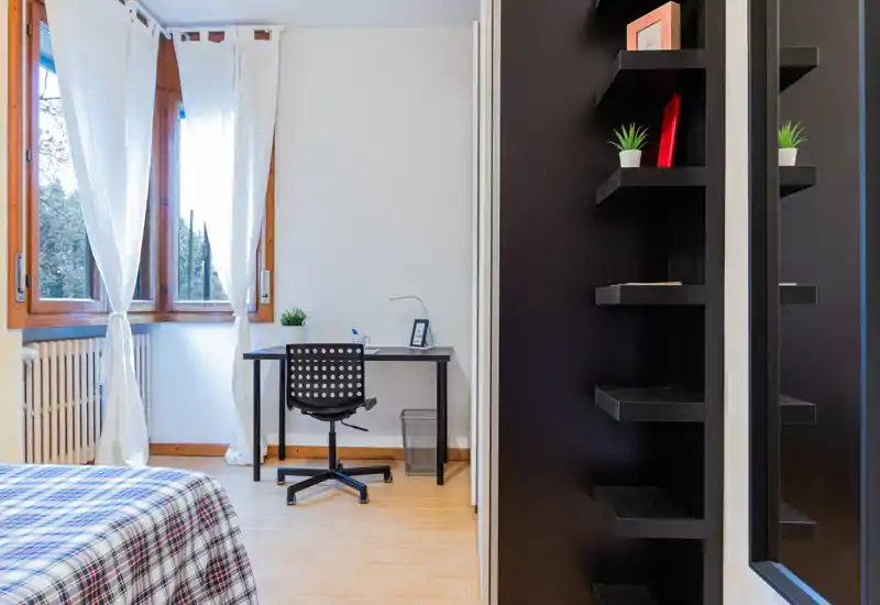 Bright, tidy bedroom with a patterned bedspread, a workspace by the window and a modern black shelving unit — ideal for remote work and comfortable stays.