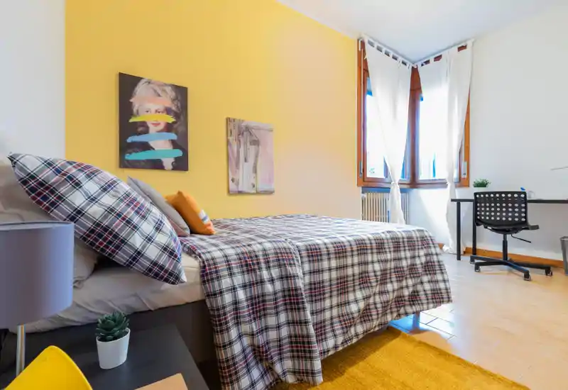 Bright, styled bedroom with a large check-pattern bed, cozy cushions and a sunny yellow accent wall. The room includes a desk by the window and warm wooden flooring, ideal for comfortable short or long stays.