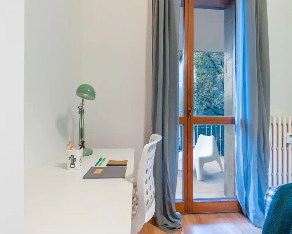 Angolo di camera con scrivania, sedia e porta finestra sul balcone. La luce naturale e le tende rendono l’ambiente luminoso e accogliente, con un accenno di biancheria che indica l’area notte vicina.
