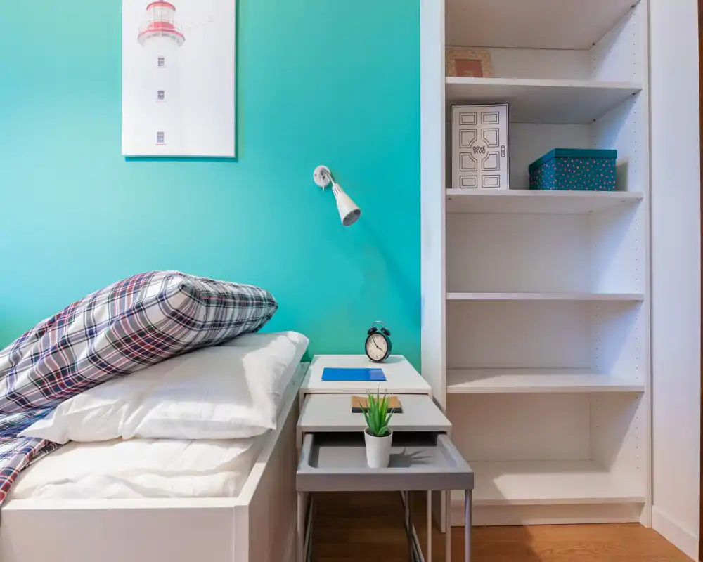 Camera da letto compatta con un letto singolo ben rifatto contro una parete color turchese, comodino con sveglia e piccola pianta e scaffalatura integrata per lo stoccaggio.