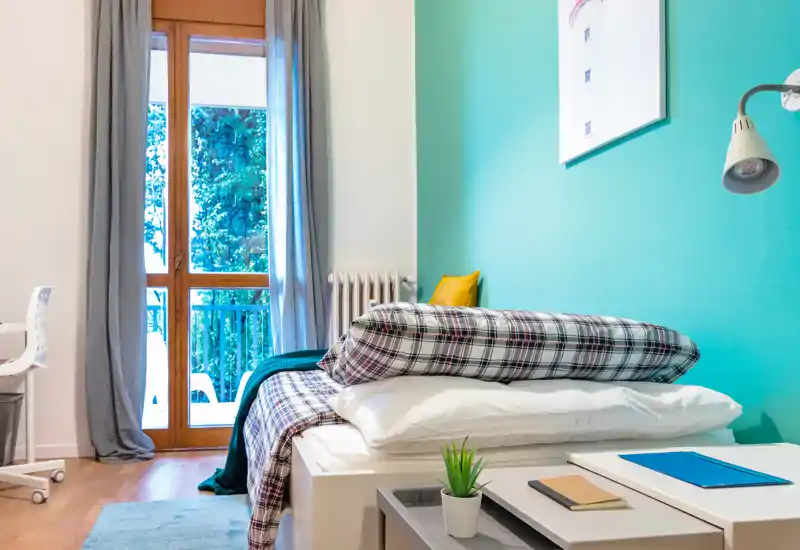 Bright, stylish bedroom with a teal accent wall, neatly made bed with plaid bedding, small bedside tables and natural light from a balcony door.
