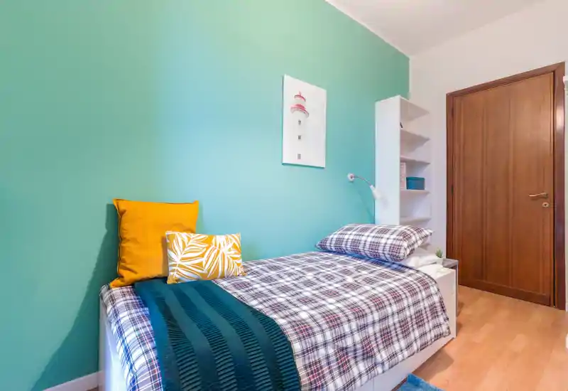 Bright, neatly styled single bedroom with a plaid bedspread, decorative pillows and a teal accent wall; compact shelving and bedside lamp complete the cozy layout.