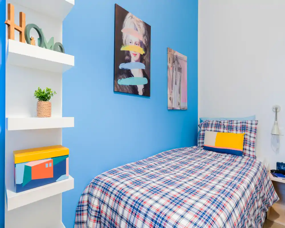 Camera da letto singola luminosa e ben arredata con copriletto a quadri, cuscino colorato e quadri moderni su una parete azzurra. Mensole compatte e lampada da comodino completano l'ambiente accogliente.
