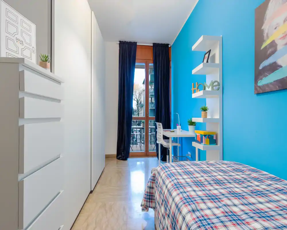Camera singola luminosa e moderna con letto a fantasia, scrivania vicino alla finestra e armadiatura integrata. La parete turchese e le mensole ordinate creano un ambiente fresco e accogliente, ideale per studenti o affitti brevi.