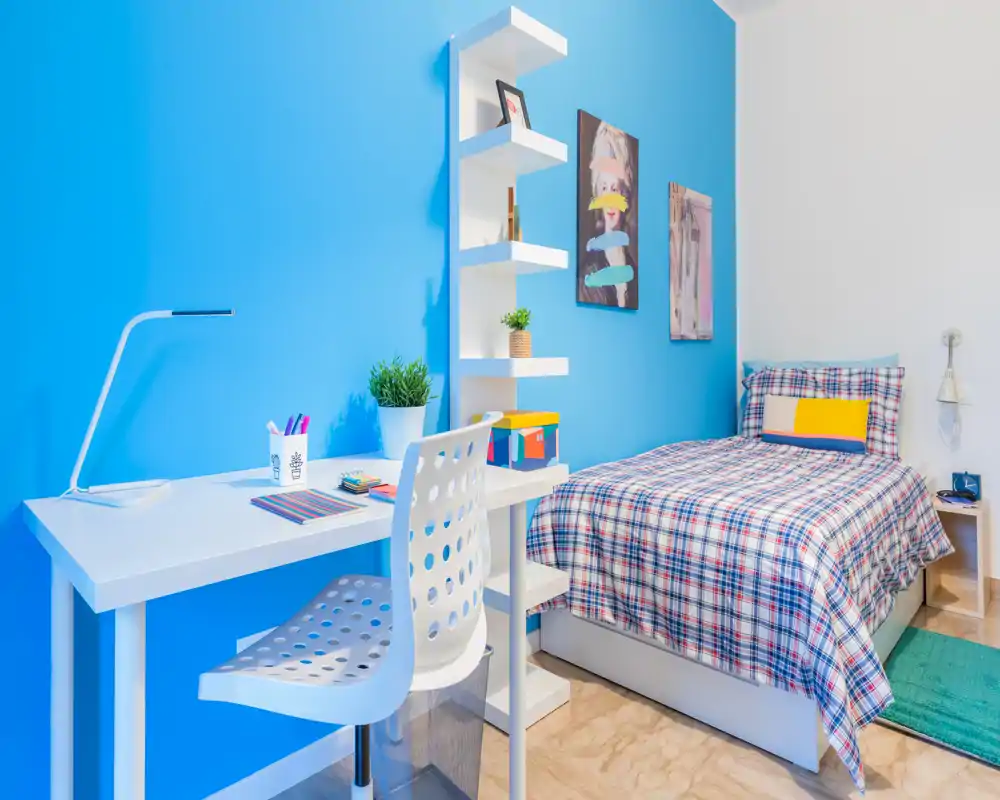 Camera da letto singola luminosa e moderna con letto in stampa scozzese ben rifatto, scrivania e scaffalatura su una parete accentata blu acceso. Linee pulite, ottima illuminazione e dettagli decorativi semplici la rendono perfetta per studenti o ospiti.