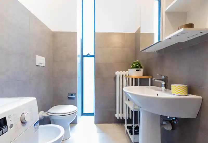 Bright, modern bathroom with a pedestal sink, toilet, bidet, radiator and a washing machine; neutral gray tiles and a tall frosted window provide natural light.
